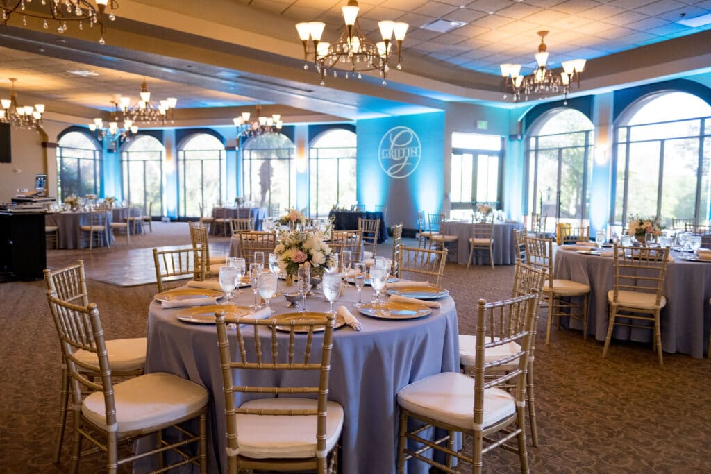 room setup for wedding reception with purple linens, gold accents, flower centerpieces, light blue uplights, and a Griffin gobo light